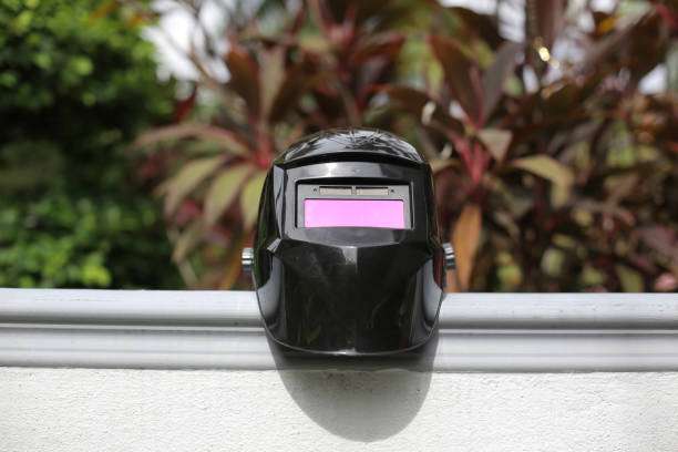 Black auto-darkening Welding Helmet with a pink visor resting on a wall, outdoors.