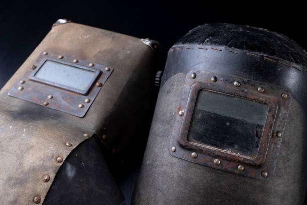 Two vintage, weathered Welding Helmets with rectangular viewports on a dark background.
