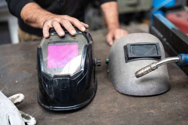 Worker's hands near two Welding Helmets: a modern auto-darkening and an old standard type.
