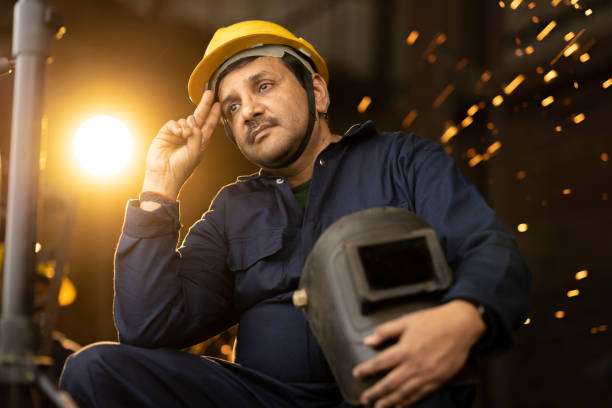 Smiling man in hard hat holding his welding helmet with sparks behind him.